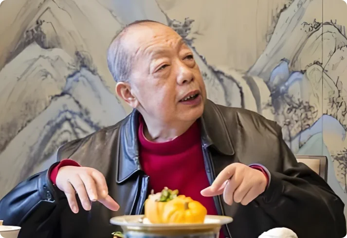 陈立先生在餐桌前，背景是水墨山水屏风，正指向前方菜肴