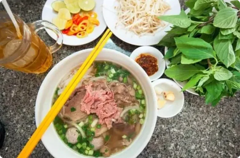 一碗越南河粉，牛肉片、肉丸配青葱，旁配豆芽、九层塔、柠檬和辣椒