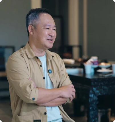 林卫辉抱臂立于茶桌前，卡其色衬衫白色T恤，背景是餐盘与茶具，神态松弛