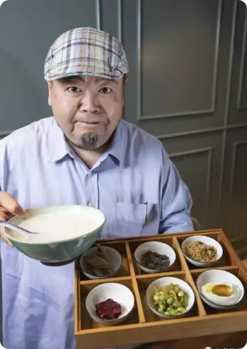 沈宏非戴格子帽、穿蓝衬衫，端着一碗白粥，面前八格小菜排列整齐