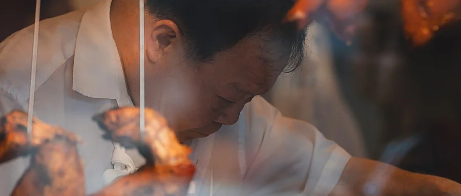 香港烧味档师傅低头斩料，头顶挂白切鸡与烧鸭，身前悬着深红叉烧